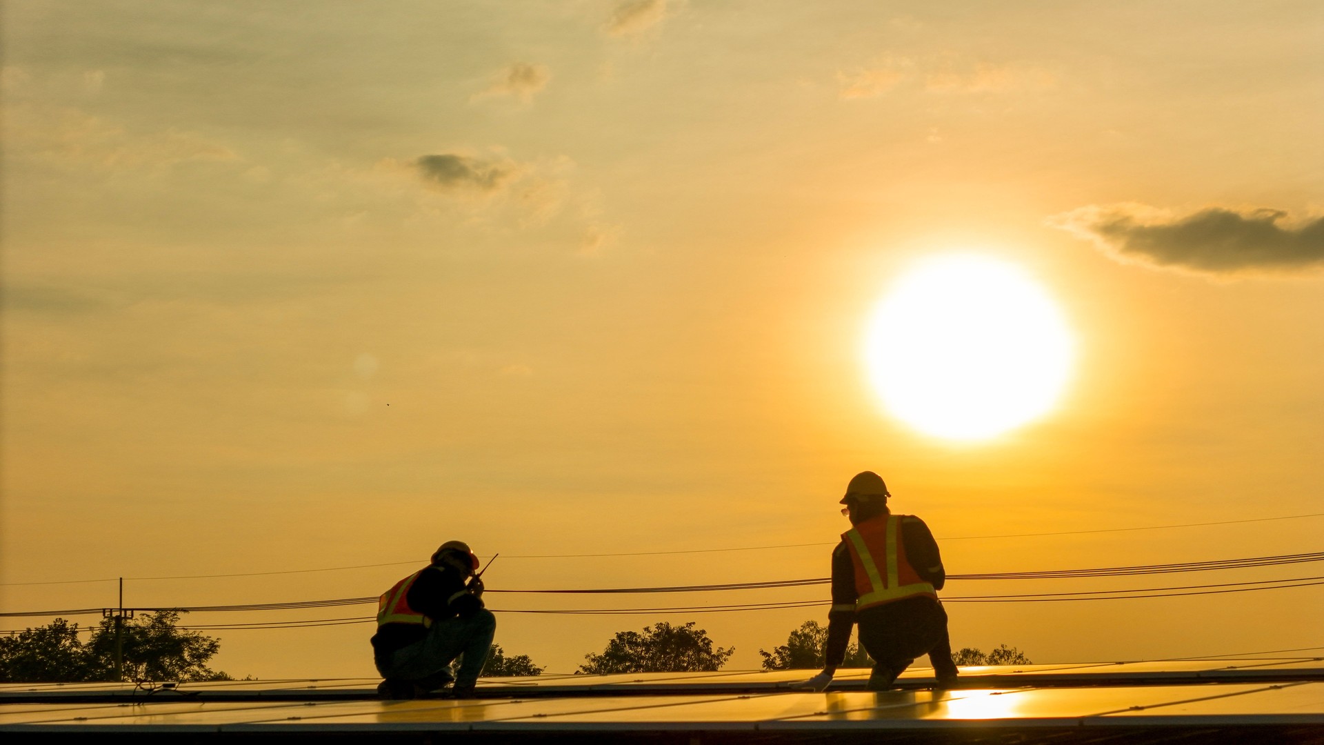 Silhouette of workers installing solar panels on a rooftop during sunset with a drone hovering nearby, highlighting renewable energy, innovation, and sustainable practices in solar technology.